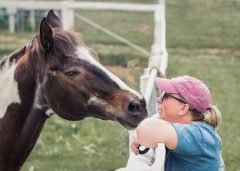 Woman leaning on fence and horse on other side of fence, sniffing her face.