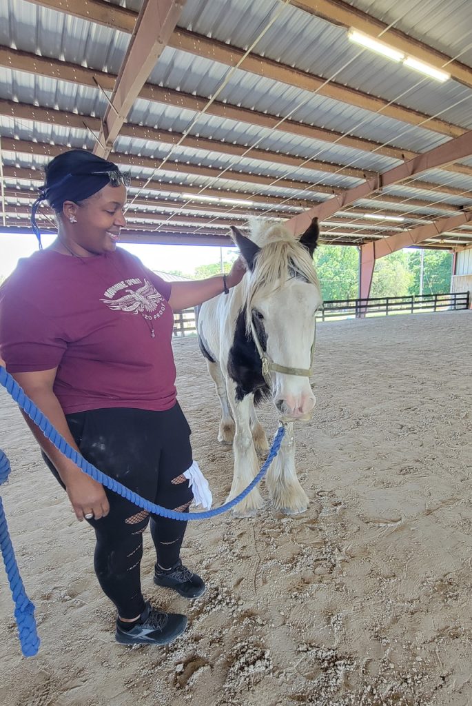VeteransBlogPost-Vet Kira Photo of woman smiling as she offers gentle touch to horse at rest.